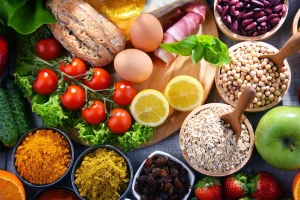 Colorful collage of fresh produce and pantry staples: tomatoes on the vine, greens, bread, eggs, lemons, apples, beans, oats, and spices on a wooden surface.