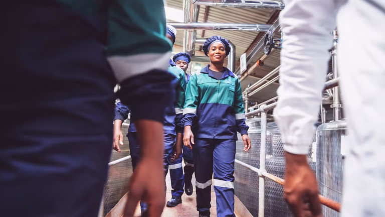 Group of factory workers in teal and navy uniforms walk down a metal walkway amid exposed pipes and railings in an industrial setting.