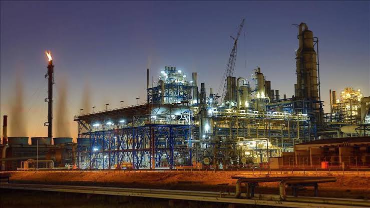 Industrial refinery complex at dusk with cranes, metal framework, and illuminated processing towers.