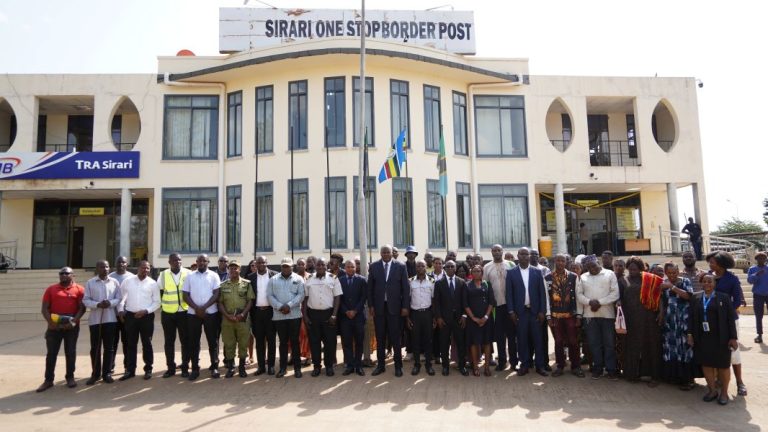 Large diverse group posing in front of the Sirari One Stop Border Post building with flags visible in the foreground.