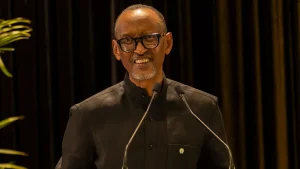 Smiling man wearing a black shirt and glasses delivering a speech at a podium with two microphones on stage.