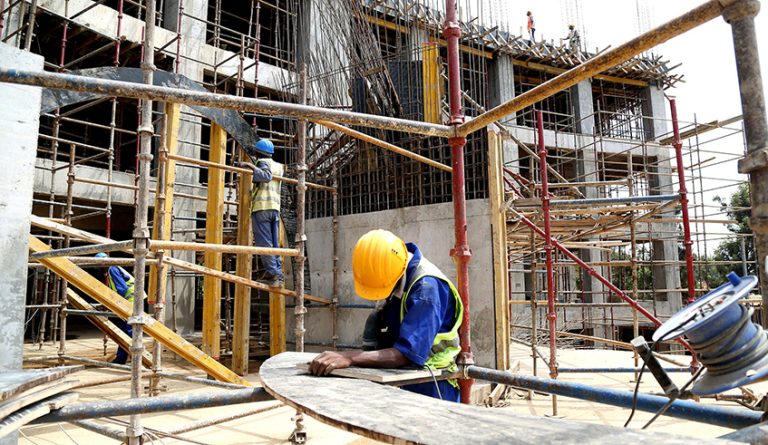 Construction site with metal scaffolding; a worker in a yellow hard hat and blue vest leans over a circular plank.